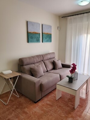 Una sala de estar con un sofá color beige, una mesita de madera con un dispositivo blanco, una mesa de centro blanca con una planta en maceta y dos cuadros abstractos en la pared.