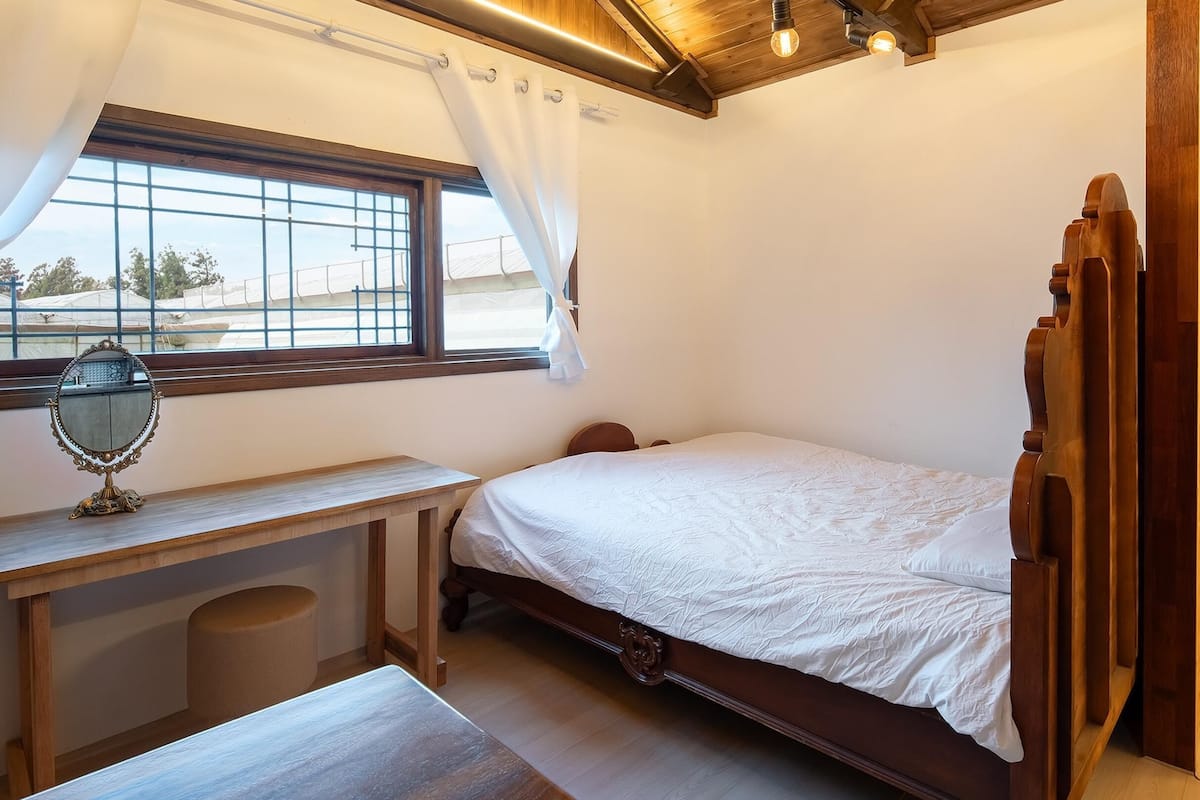 Ein Schlafzimmer mit einem hölzernen Bett, einem Nachttisch mit Spiegel, einem Fenster mit Vorhängen und Blick ins Freie.