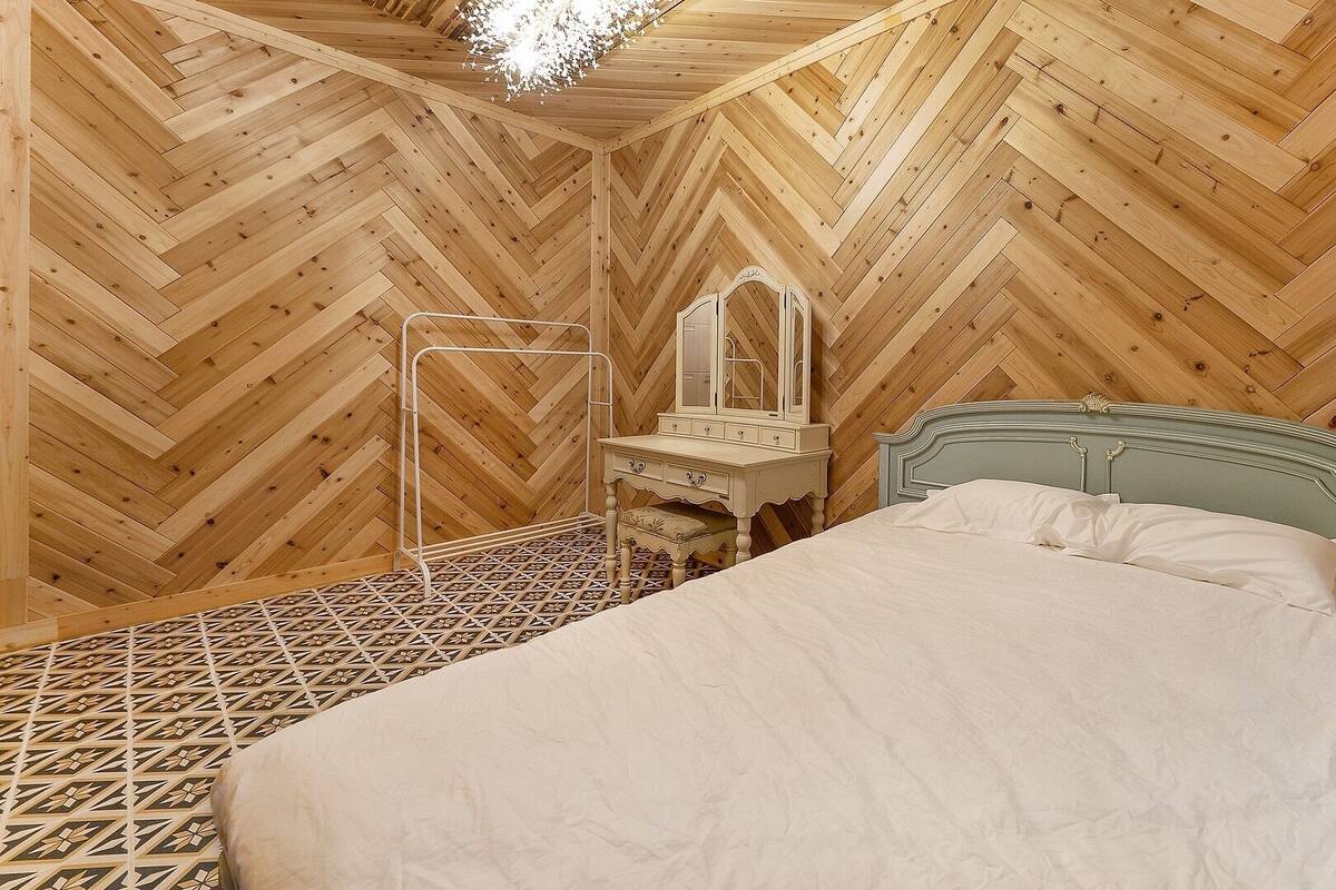 Ein Schlafzimmer mit Fischgrätparkett, einem Bett mit gemustertem Kopfteil, einem Schminktisch mit Spiegel und einer Hängeleuchte.