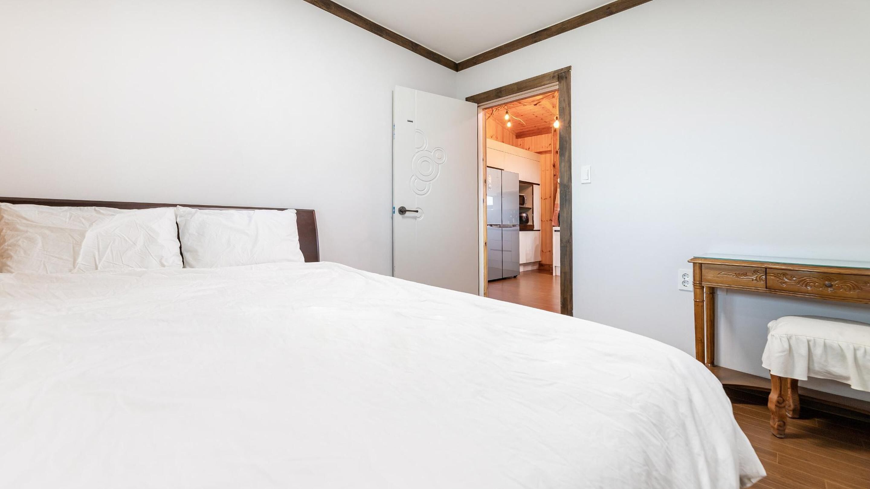 A bedroom with a large bed, a wooden desk, and a door leading to a kitchen area.