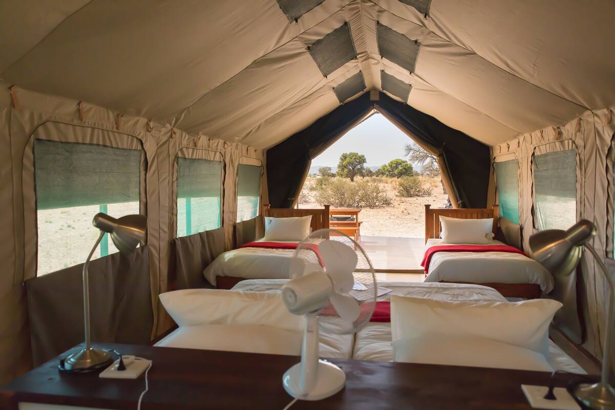 A tented accommodation with two beds, each with a pillow and a blanket, and a table with a lamp.