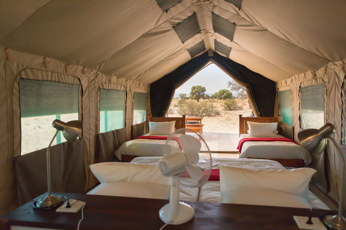 A tent with two beds, each with a pillow and a blanket, a table with a lamp, and a view of the outdoors.