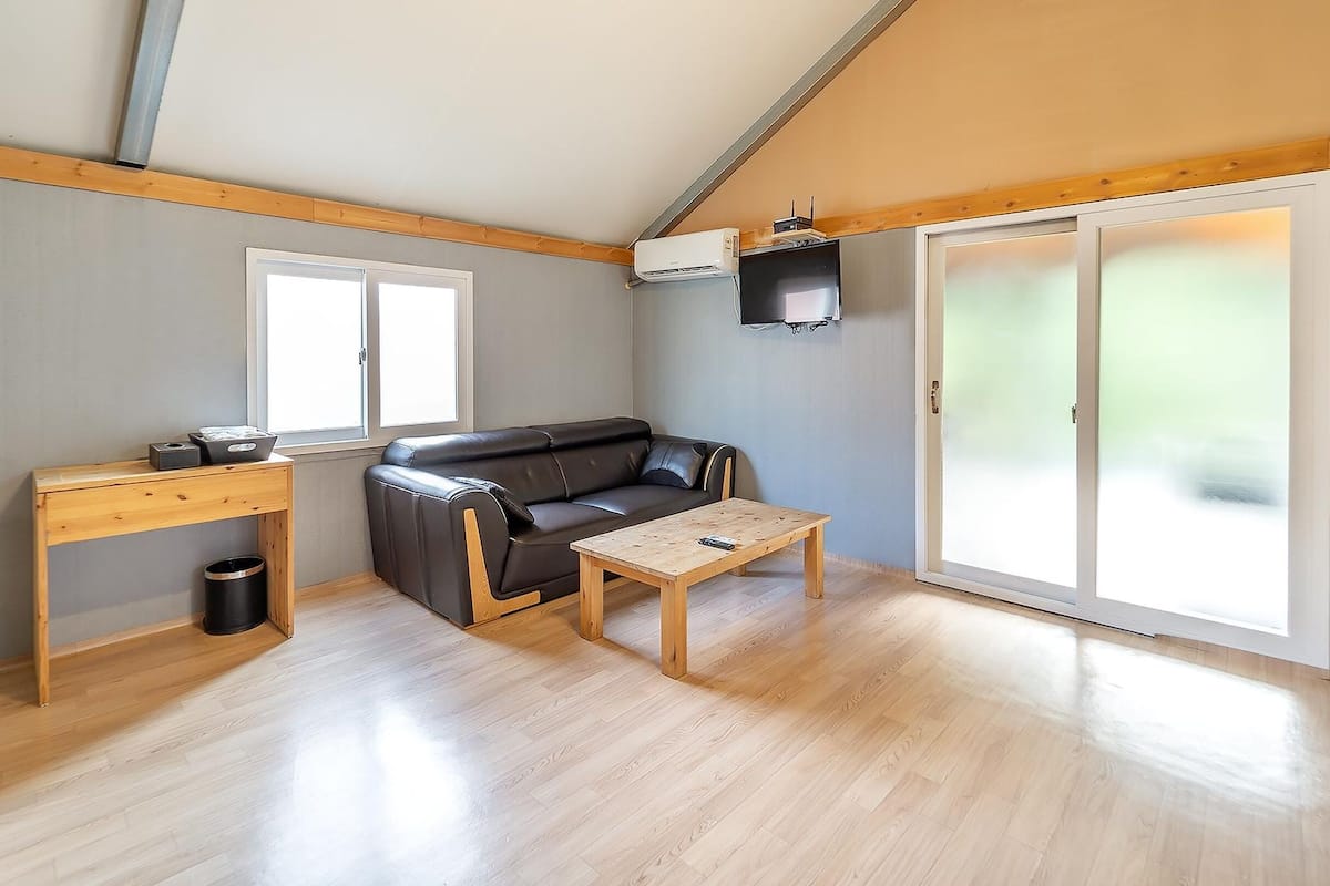 A compact living space with a wooden floor, a sofa, a small table, a desk, and a sliding glass door.