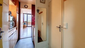 Un pasillo estrecho con armarios de madera y un microondas a la izquierda, una puerta con espejo a la derecha y vistas a una habitación con escritorio y silla.
