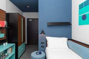 Una moderna camera d'albergo con un letto, una scrivania con una sedia, un tavolino con una lampada e una mensola a parete.