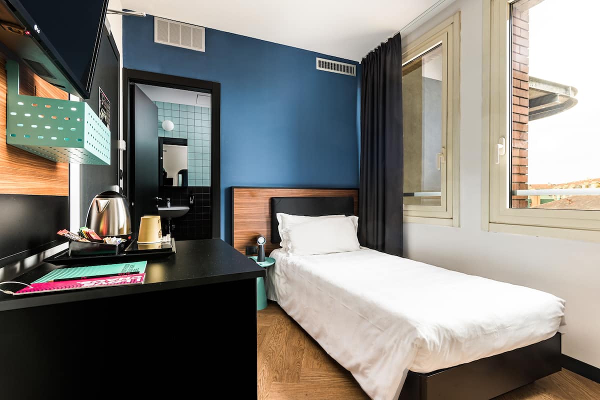 A hotel room with a bed, a desk with a kettle, a cup, and a notebook.