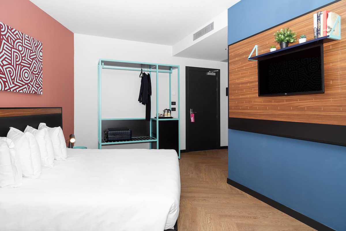 A modern hotel room with a bed, a teal wardrobe, a TV mounted on a wooden wall, and a patterned wall art piece.