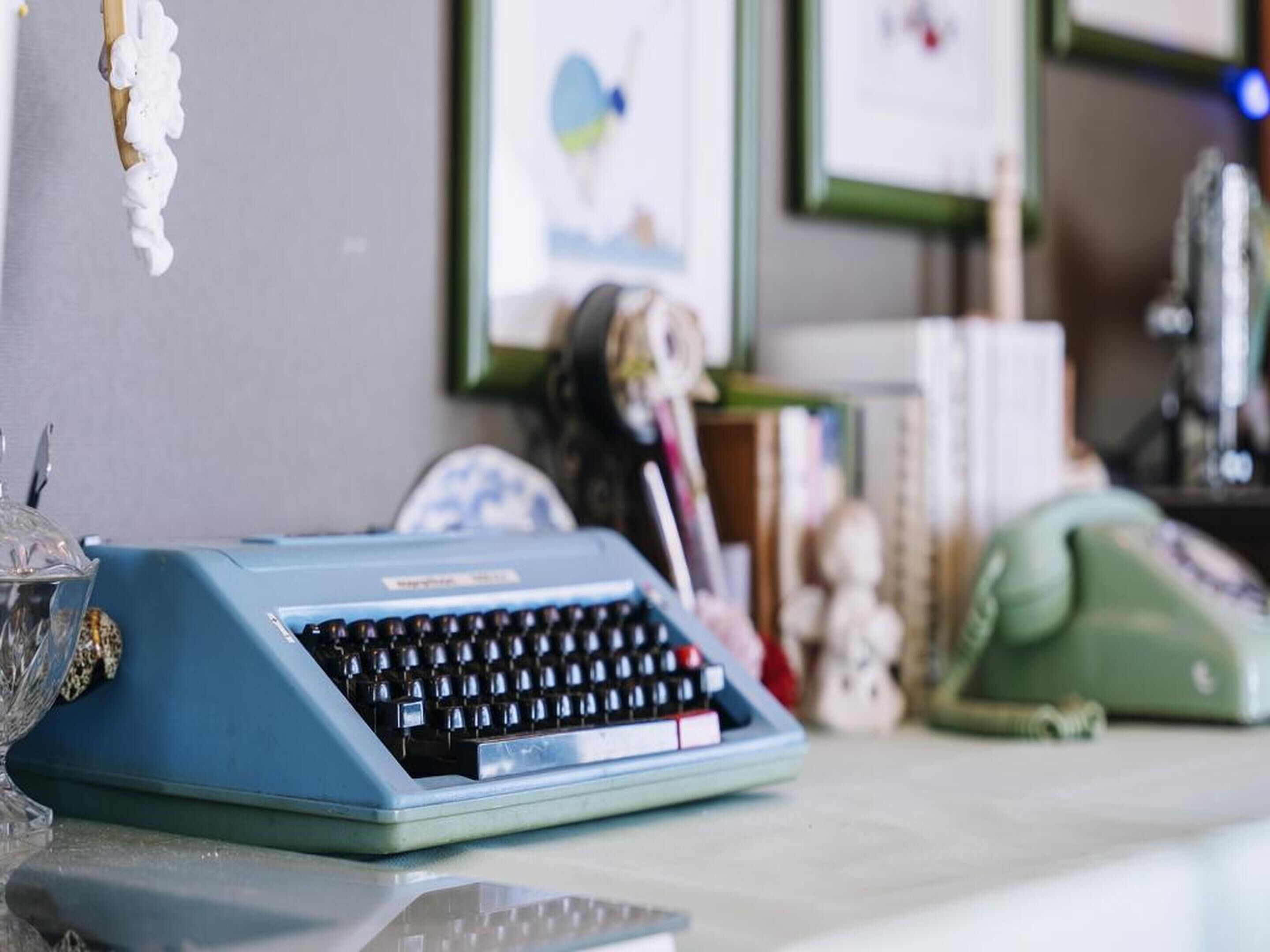 A vintage blue typewriter on a desk with a glass vase, a green telephone, and framed pictures on the wall.