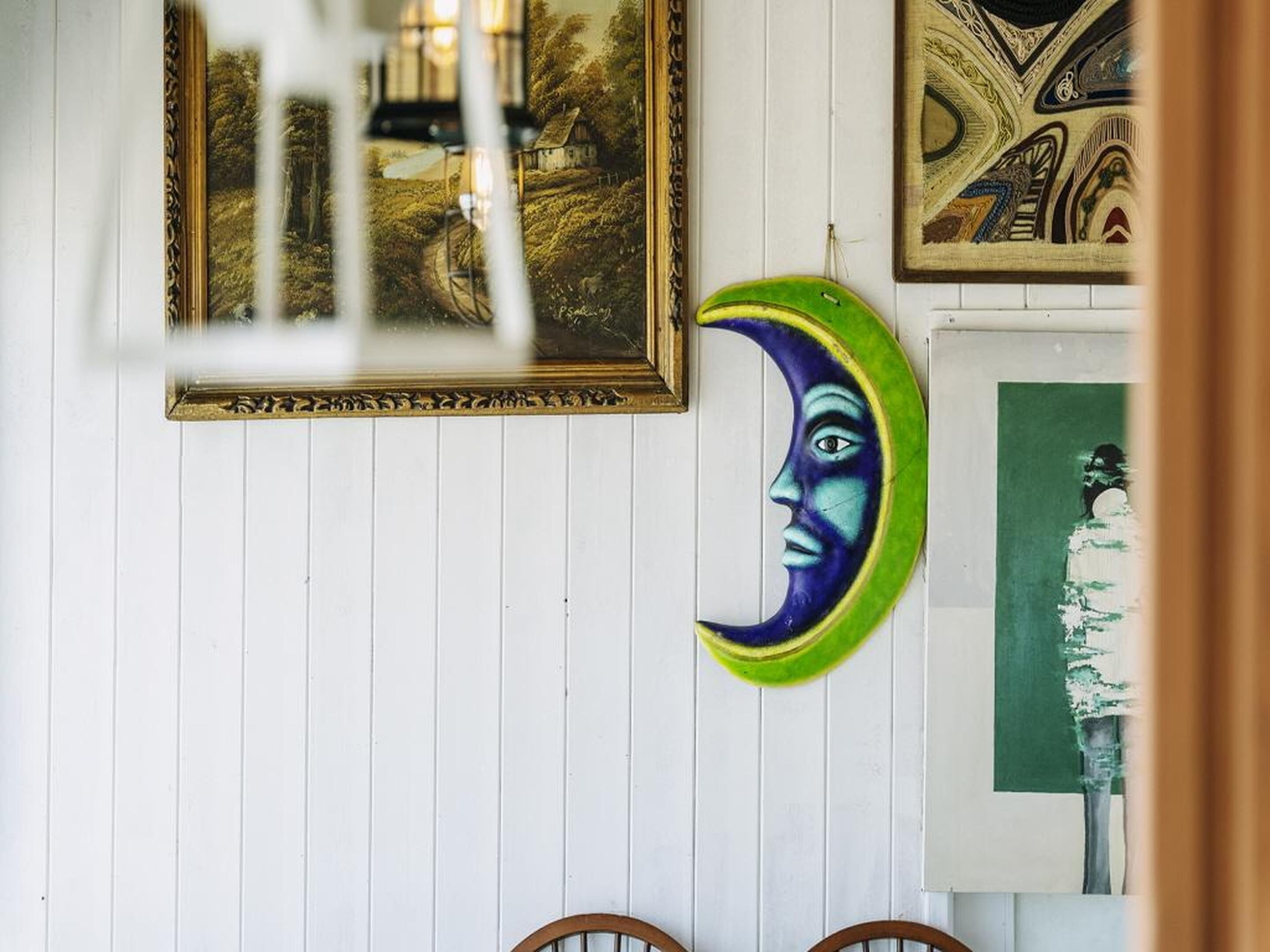 A room with a white wooden wall, a framed painting, a green and blue crescent moon decoration, and a green abstract painting.