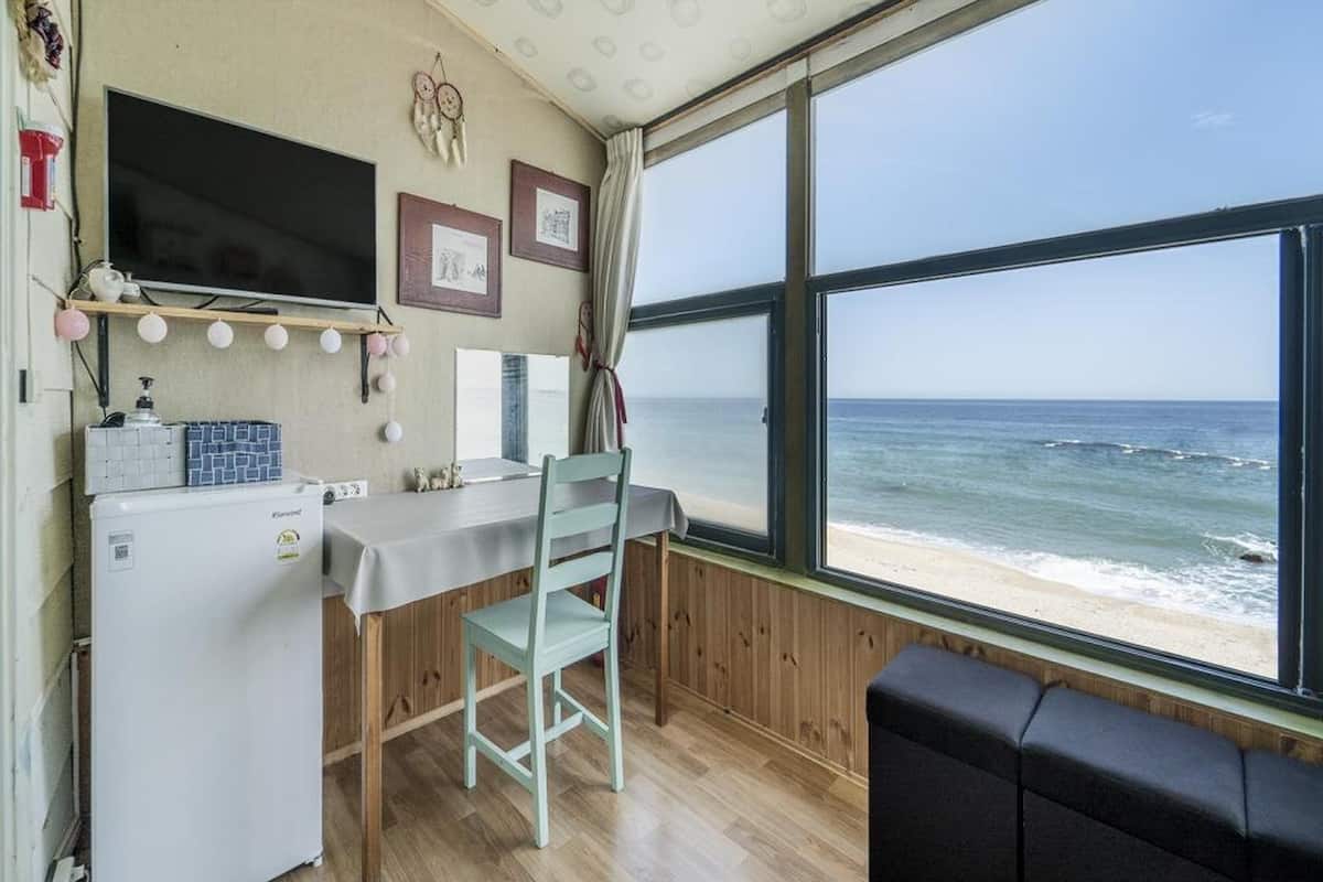 Ett litet rum med trägolv, ett litet bord, en stol, en soffa, en tv och ett fönster med utsikt över stranden.
