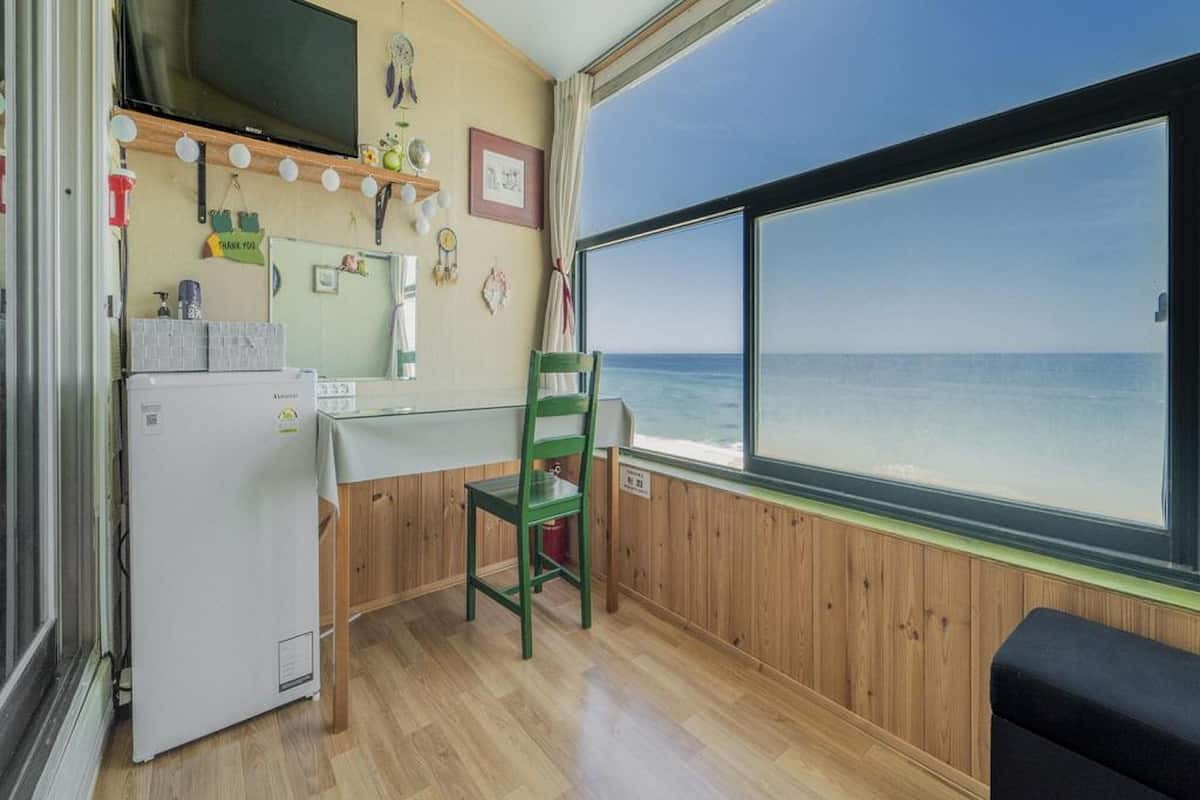 Ett kompakt kök med ett kylskåp, ett matbord i trä och en stol. Ett stort fönster ger utsikt över havet.