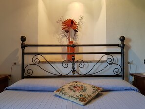 Een bed met een metalen hoofdbord, een bloemenkussen en een decoratieve vaas met gedroogde bloemen op een muur.