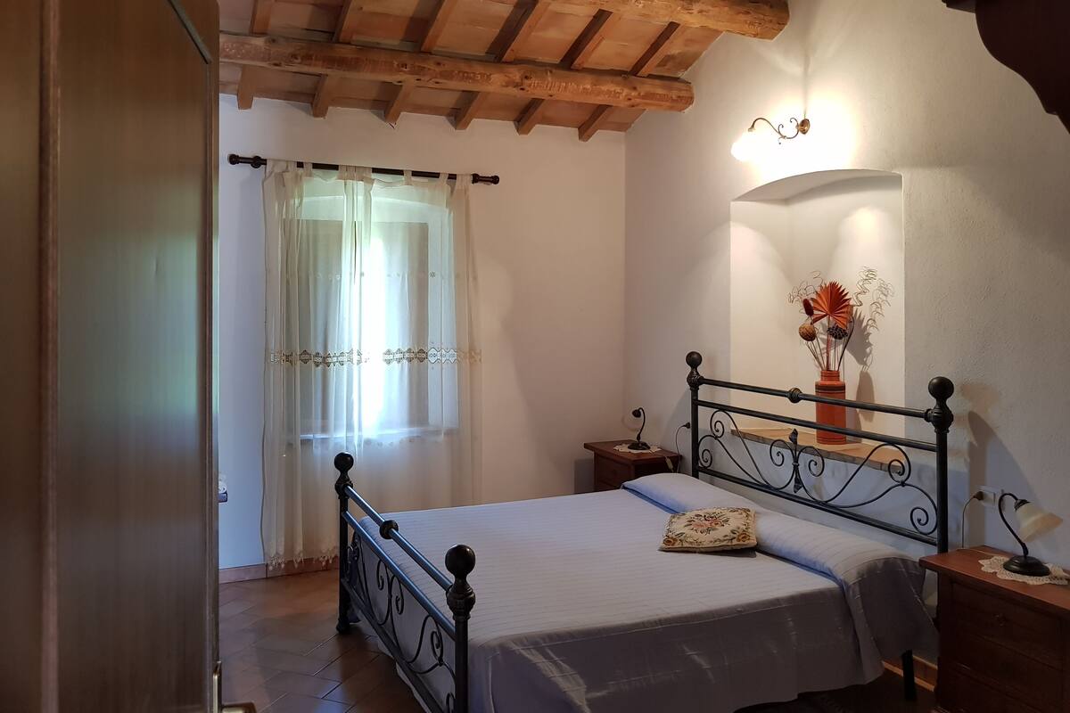 Une chambre avec un plafond en bois, un lit avec un cadre en métal, une table de chevet, un vase avec des fleurs et une lampe fixée au mur.