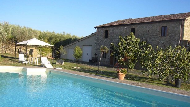 A swimming pool with a clear blue water surface, surrounded by a stone building with a tiled roof, a covered seating area, and potted plants.