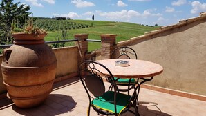 A terrace with a round table and chairs, a large terracotta pot, and a view of a green hillside.