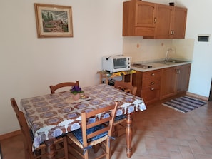 Een compacte keuken met houten kastjes, een spoelbak en een broodrooster. Er is een tafel met stoelen en een lijst met een foto aan de muur.