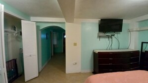 Un dormitorio con cama, cómoda, televisor montado en la pared y un pasillo que conduce a otra habitación.