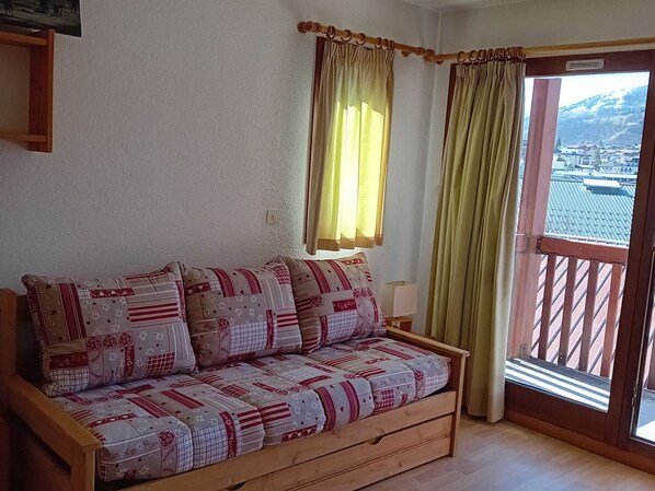 Ein Holzsofa mit gemusterten Kissen, ein kleiner Tisch mit einer Lampe und Blick auf eine gebirgige Landschaft durch ein Fenster.
