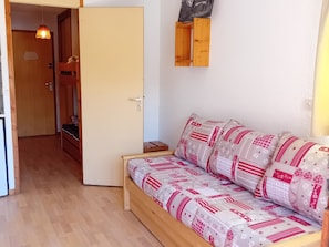 Ein Zimmer mit einer hölzernen Bank, einem Bett mit gemusterten Textilien, einem Holzschrank und einer Tür zu einem anderen Raum.
