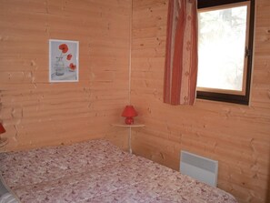 A wooden cabin room with a bed, a red lamp, a window with curtains, and a framed picture on the wall. - 1-Bed Apartment with Balcony, Fully Equipped Kitchen, and Mountain Views (Bolquère-Pyrénées 2000)