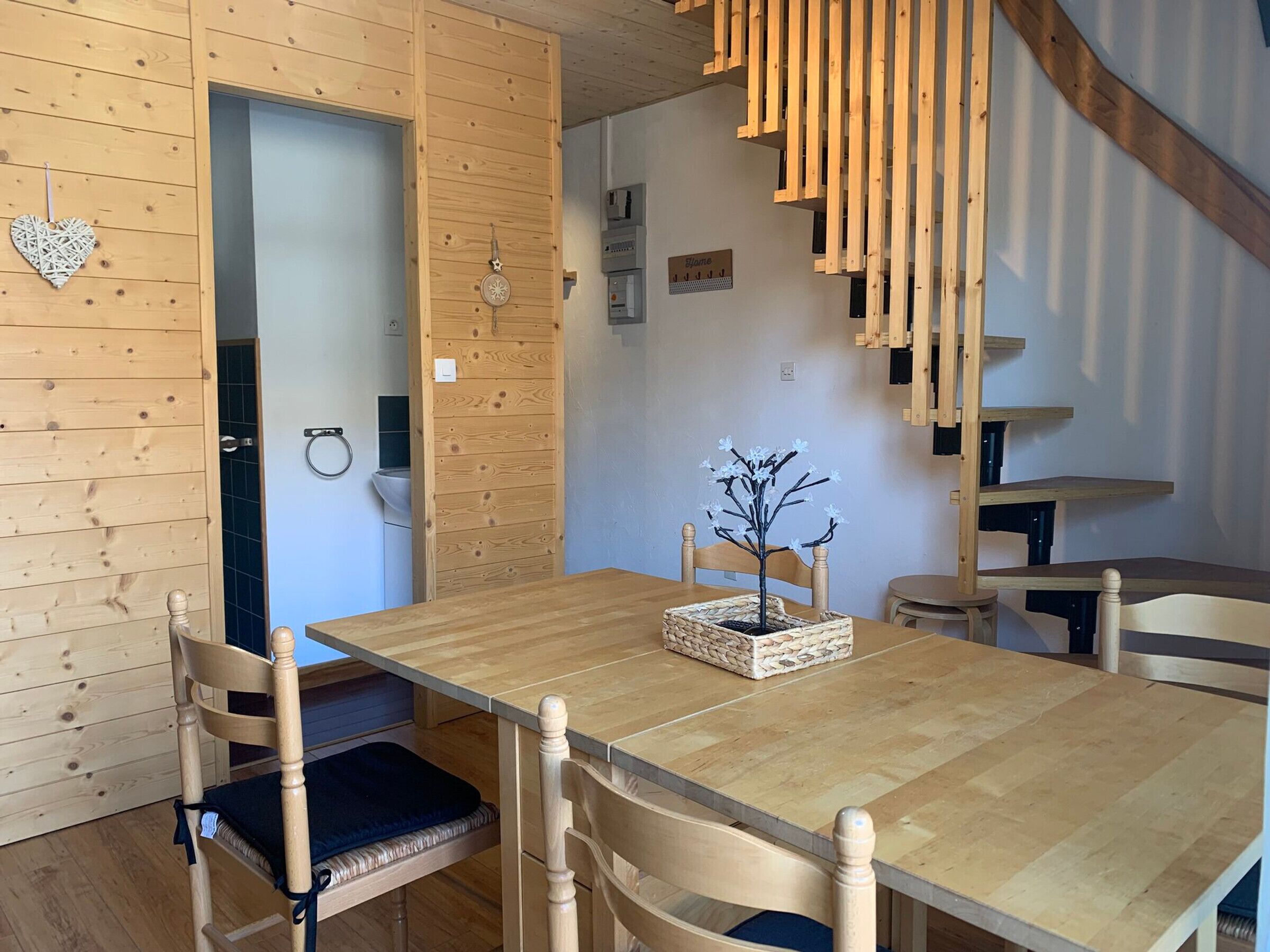 A wooden interior with a dining table, chairs, a small decorative tree, and a staircase.