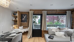 A modern living room with a dining area, wooden accents, a large window with curtains, and a view of greenery outside. - Charming 3-BR Apartment in Megève, Near Center, Ski, and Sports Complex (Megève)