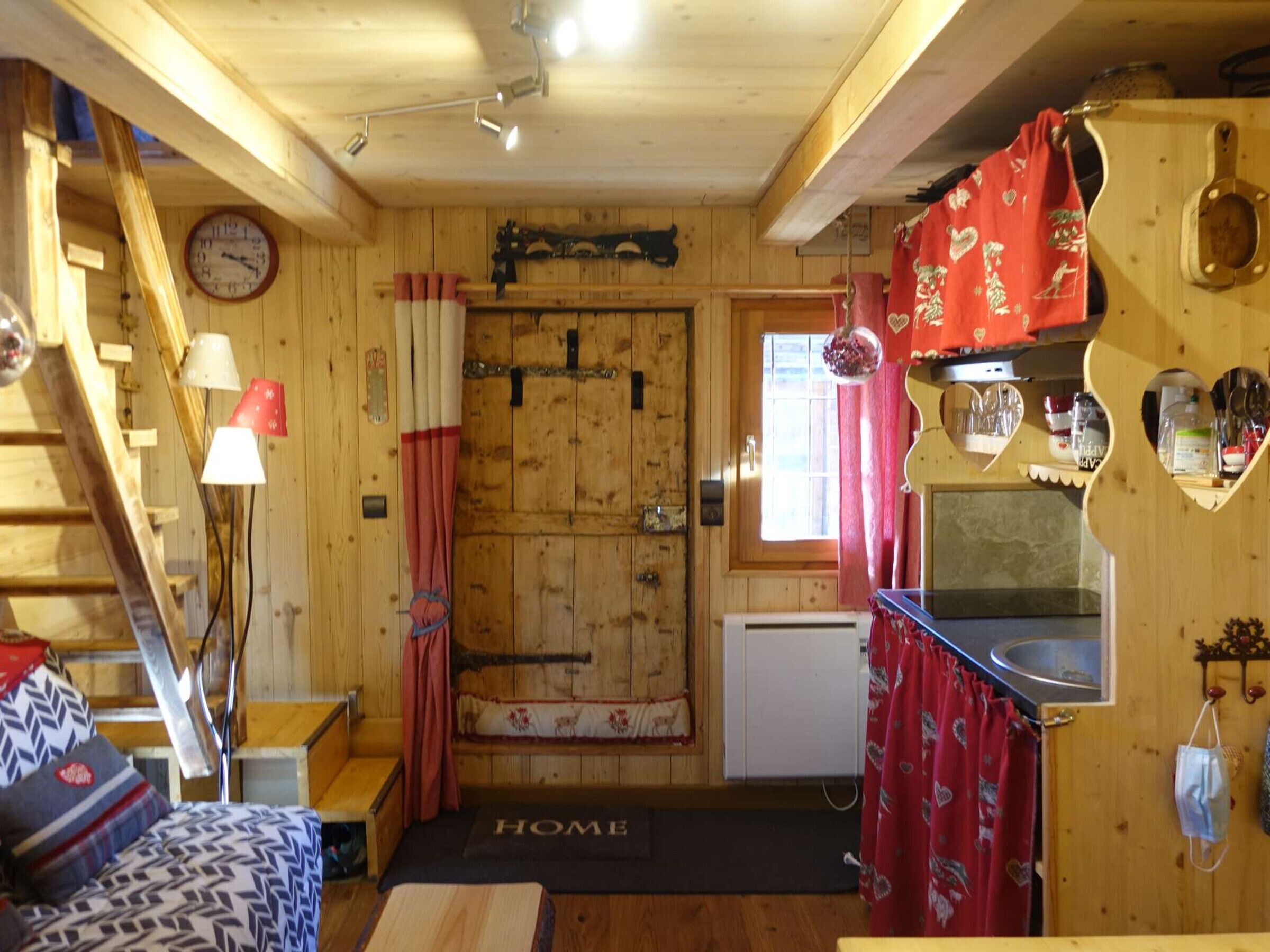 Interior acolhedor de uma cabana com decoração em madeira, um relógio na parede, um pequeno espaço de cozinha e uma escada.
