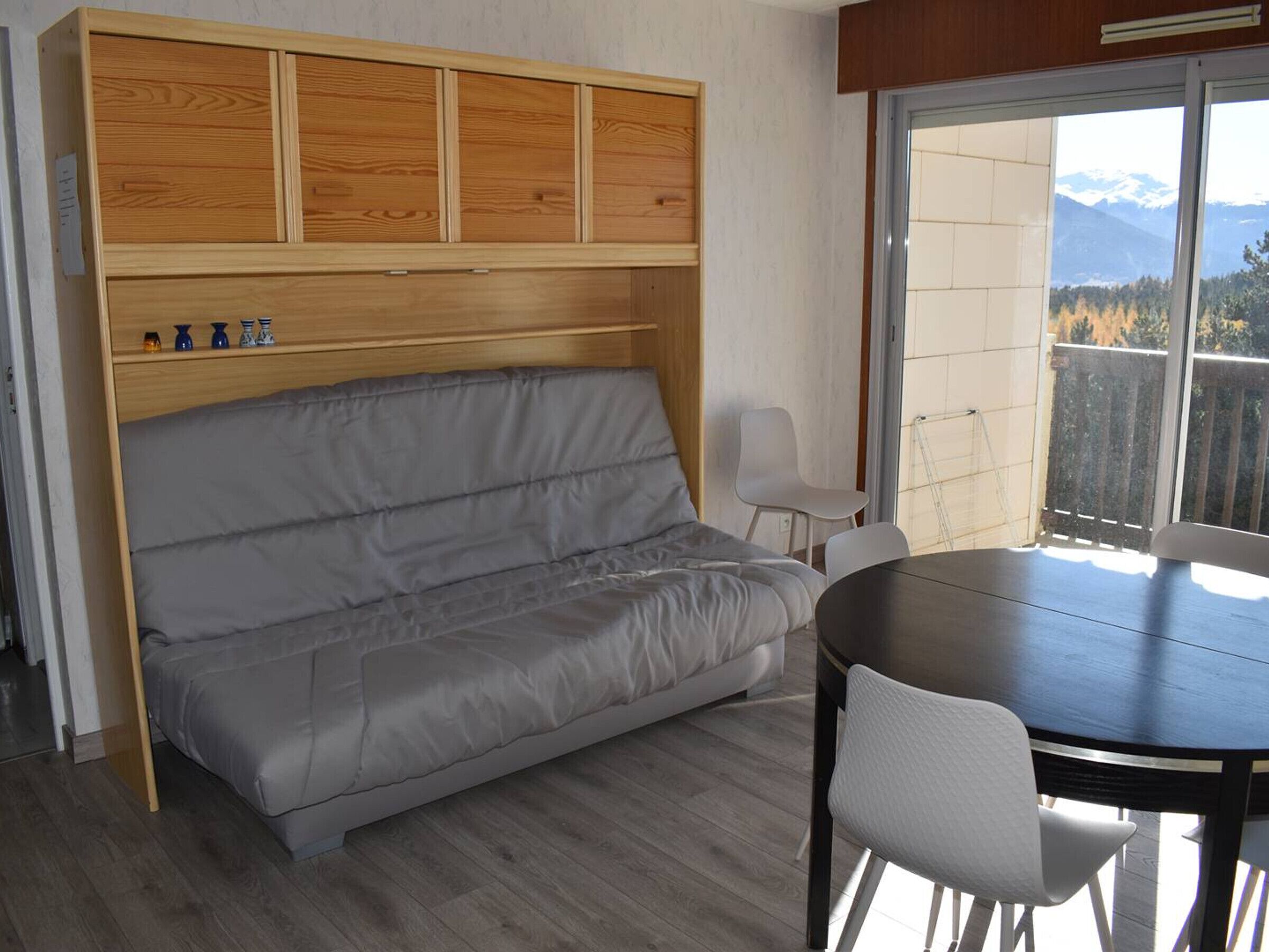 Quarto compacto com sofá embutido, mesa de jantar e vista para montanhas através de uma janela ampla.