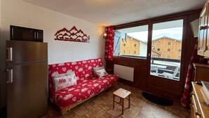 Un espacio habitable compacto con kitchenette, un sofá con ropa de cama estampada, una pequeña mesa de madera y vistas a edificios a través de una ventana.