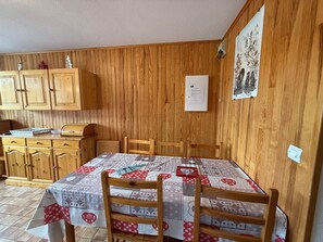 A dining area with a wooden wall, a table with a quilted tablecloth, wooden chairs, and a wooden cabinet.
