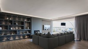 Ein modernes Wohnzimmer mit einer Eckcouch, einer dunklen Regaleinheit, einem Flachbildfernseher und Blick auf Boote durch große Fenster.