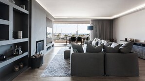Ein modernes Wohnzimmer mit grauem Sofa, Fernseher und Bücherregal. Ein Balkon bietet Blick auf Boote, und ein Essbereich mit Tisch und Stühlen lädt zum Verweilen ein.