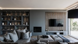 Ein modernes Wohnzimmer mit grauem Sofa, einem Flachbildfernseher an einer Holzpaneelwand und integrierten Regalen mit verschiedenen Dekorationselementen.