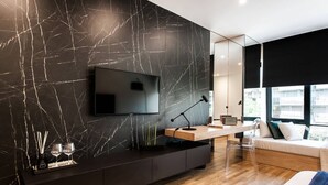 A modern bedroom with a large flat-screen TV mounted on a marble wall, a bedside table with a lamp, a sofa with cushions, and a window with a city view. - Executive Luxury Suite Athens Riviera by The Olon (Glyfada)
