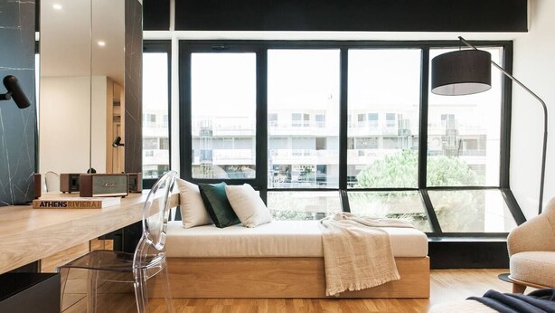 A modern bedroom with a wooden floor, a bed with pillows, a desk with a lamp, and large windows offering a city view. - Executive Luxury Suite Athens Riviera by The Olon (Glyfada)