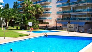 Zona de piscina con un puesto de salvavidas, palmeras y un edificio de varios pisos con balcones.