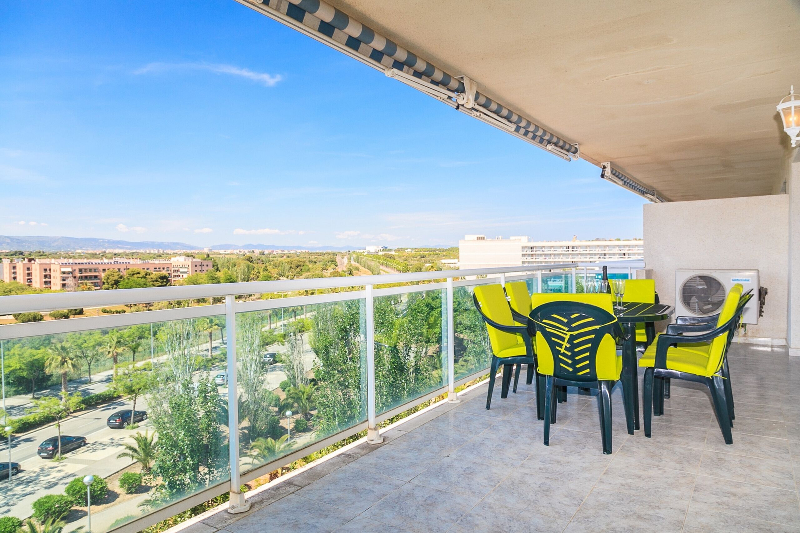 Un balcón con sillas amarillas y una mesa, con vistas a un paisaje urbano con edificios y zonas verdes.