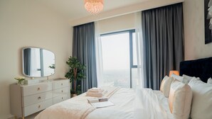 A modern bedroom with a large bed, a dresser, a mirror, and a view of the city. - Nasma Luxury Stays - Dubai Hills (Dubai)