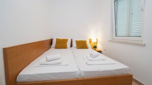 A double bed with white linens and two yellow pillows, a bedside lamp, and a window with white shutters. - Apartments Adela II (Novalja)