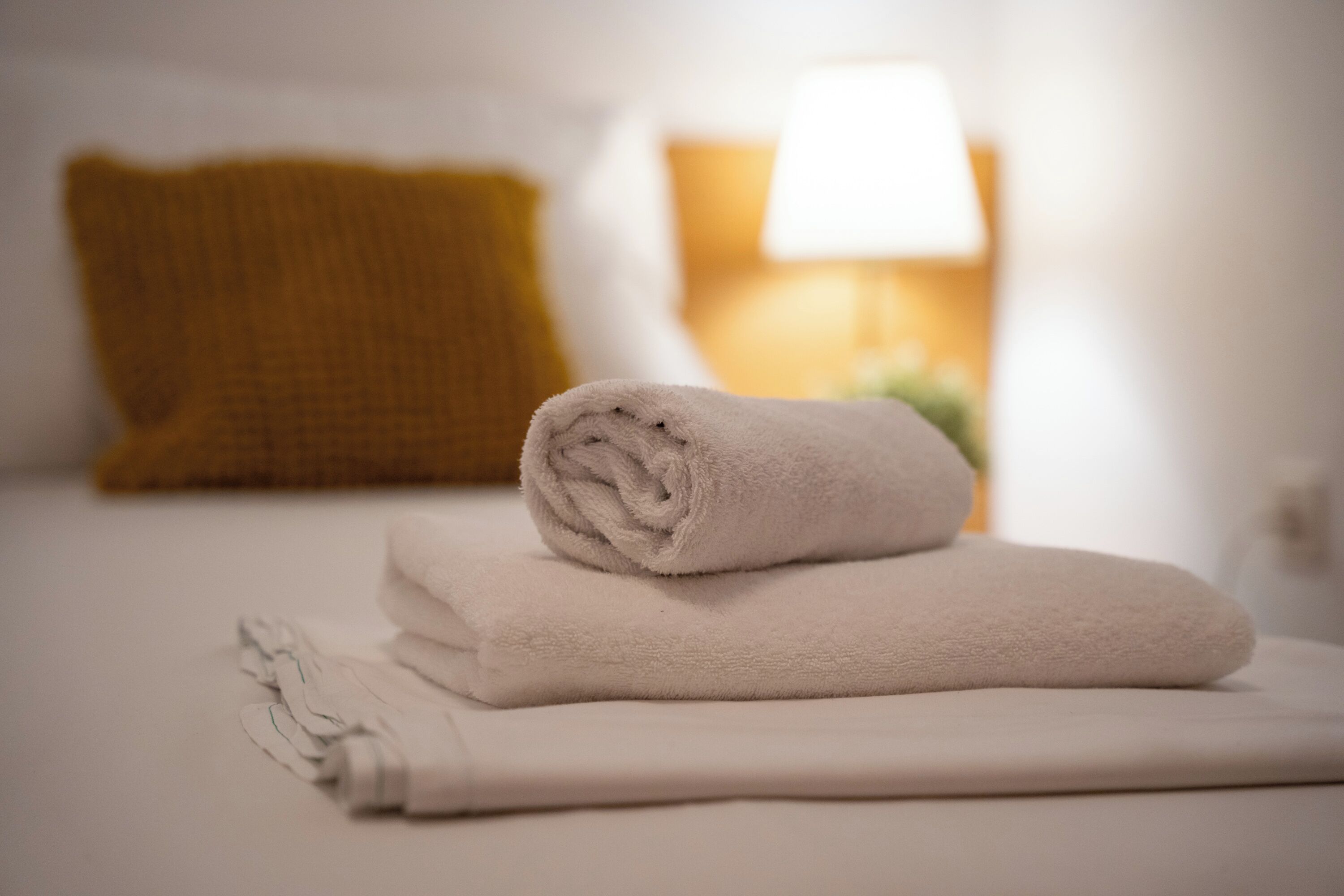 Ein ordentlich bezogenes Bett mit einem aufgerollten weißen Handtuch und einem gefalteten weißen Handtuch darauf, einem senfgelben Kissen und einer Nachttischlampe mit weißem Lampenschirm.