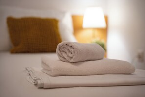 Una cama bien hecha con una toalla blanca enrollada y otra doblada encima, una almohada color mostaza, y una lámpara de noche con pantalla blanca.