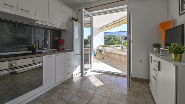 A modern kitchen with white cabinets, a built-in oven, and a sliding glass door leading to an outdoor patio with a table and chairs. - Apartments Adela II (Novalja)