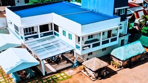Un bâtiment moderne à deux étages, doté d’un toit plat, d’un espace extérieur couvert aménagé en coin salon et d’une structure adjacente avec un toit de chaume.