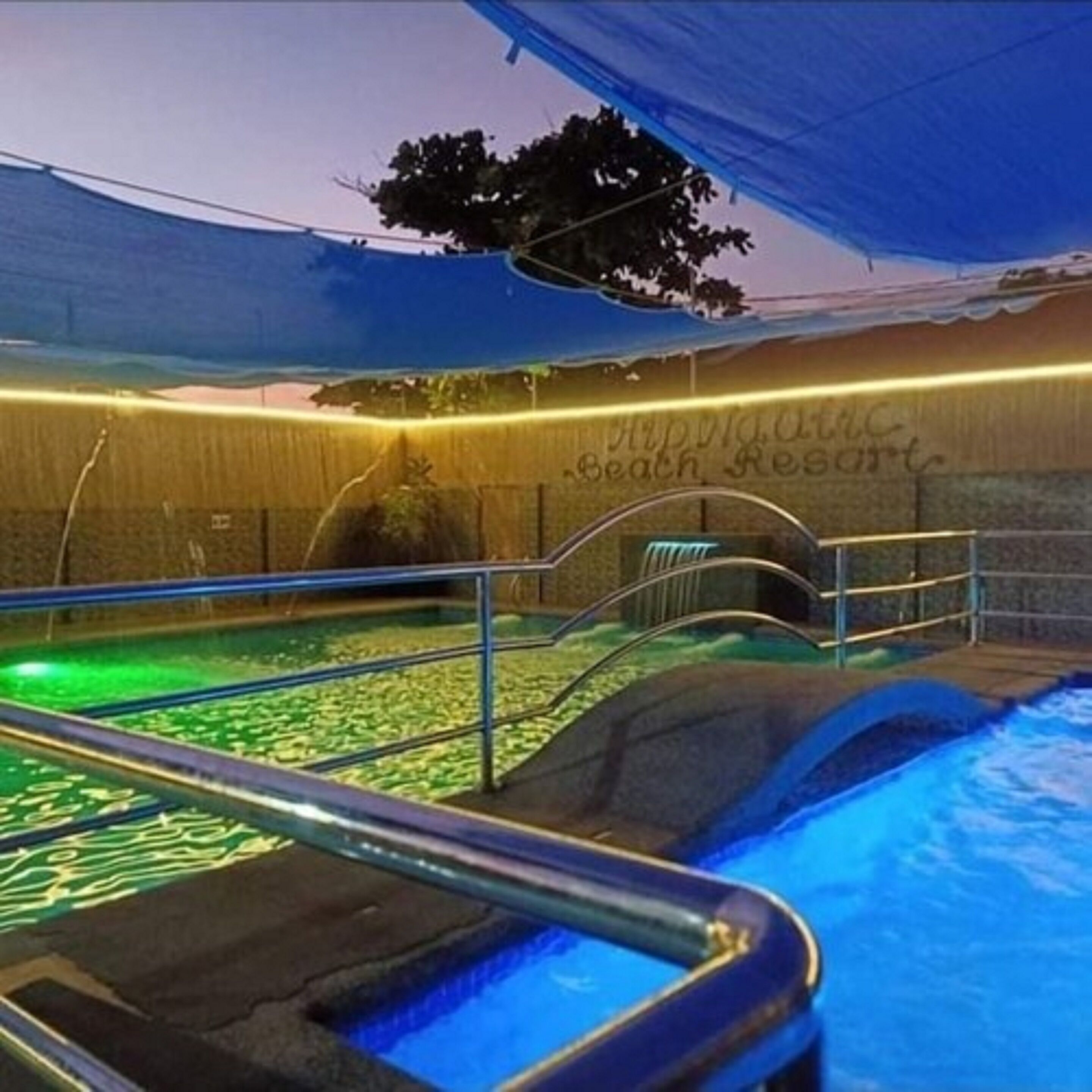 A swimming pool with a blue water feature, surrounded by a wooden deck and a covered area with a sign reading "Beach Resort".