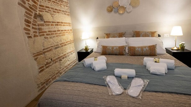 Een hotelkamer met een bakstenen muur, een bed met meerdere kussens en een blauwe plaid, twee nachtkastjes met lampen, en netjes opgemaakte handdoeken op het bed.