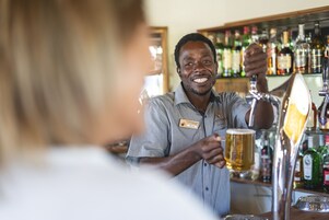 Um barman servindo uma cerveja num bar, com prateleiras cheias de garrafas de bebidas alcoólicas ao fundo.