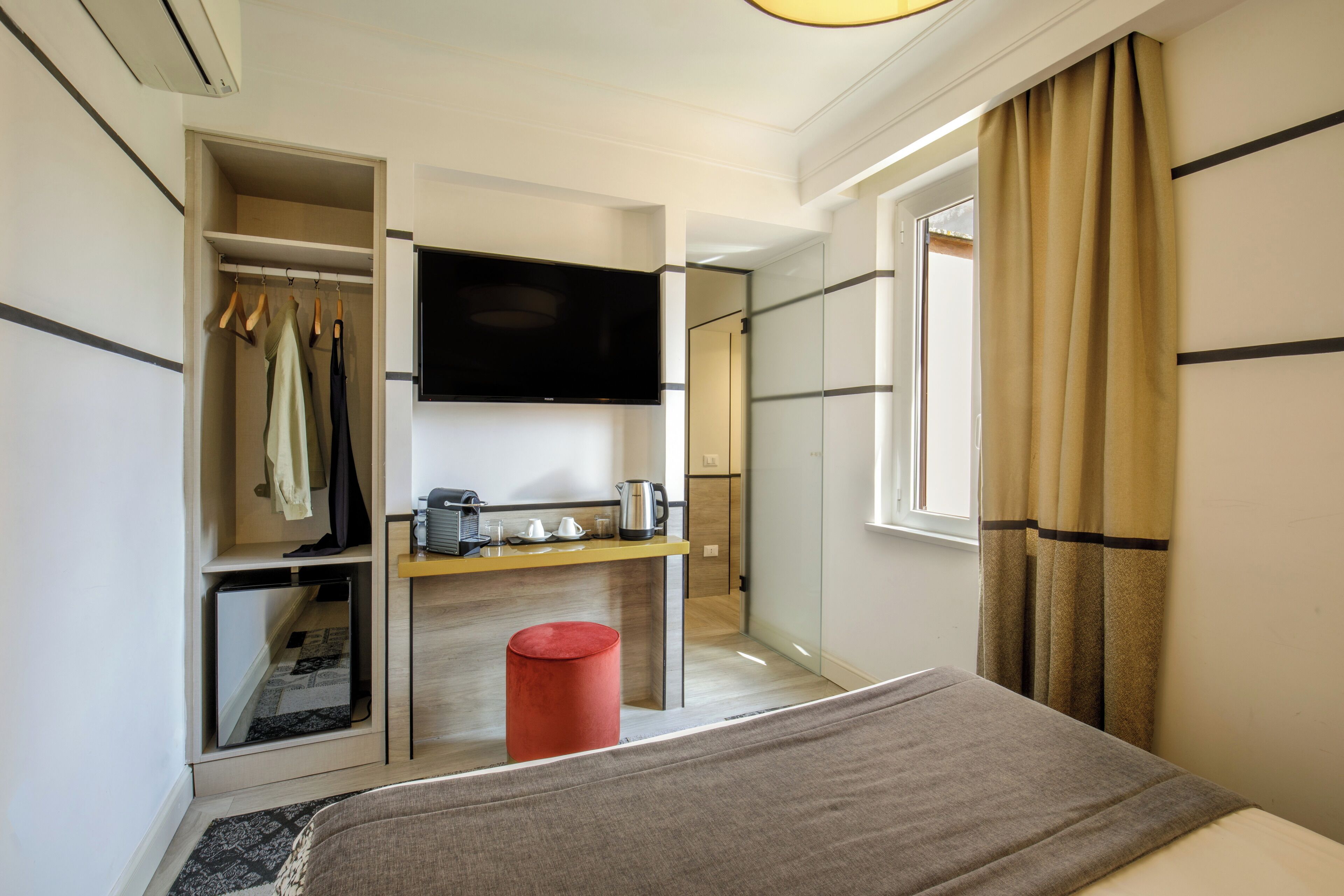 Una habitación de hotel moderna con cama, televisión montada en la pared, una pequeña kitchenette y un armario con ropa colgada.