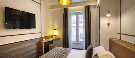 A modern hotel room with a large bed, a flat-screen TV, a bedside table, a lamp, and a view of the outside through a window.