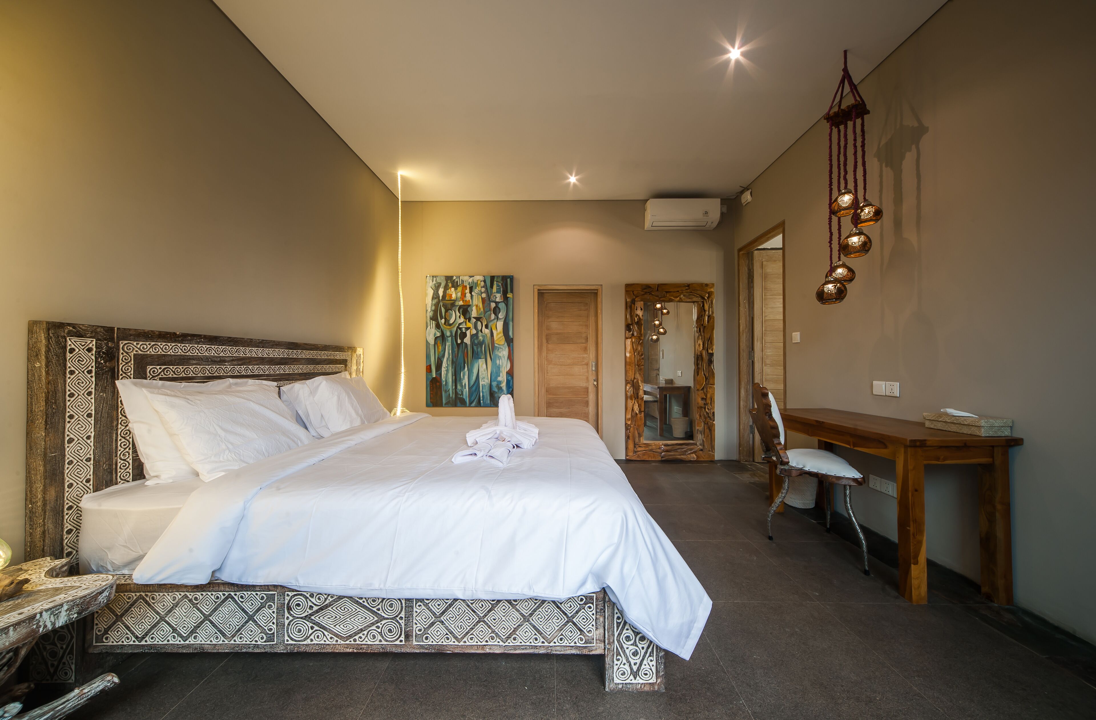 Una cama bien hecha con sábanas blancas, cabecera de madera y piecero estampado. Hay un escritorio de madera con una silla y una lámpara colgante.