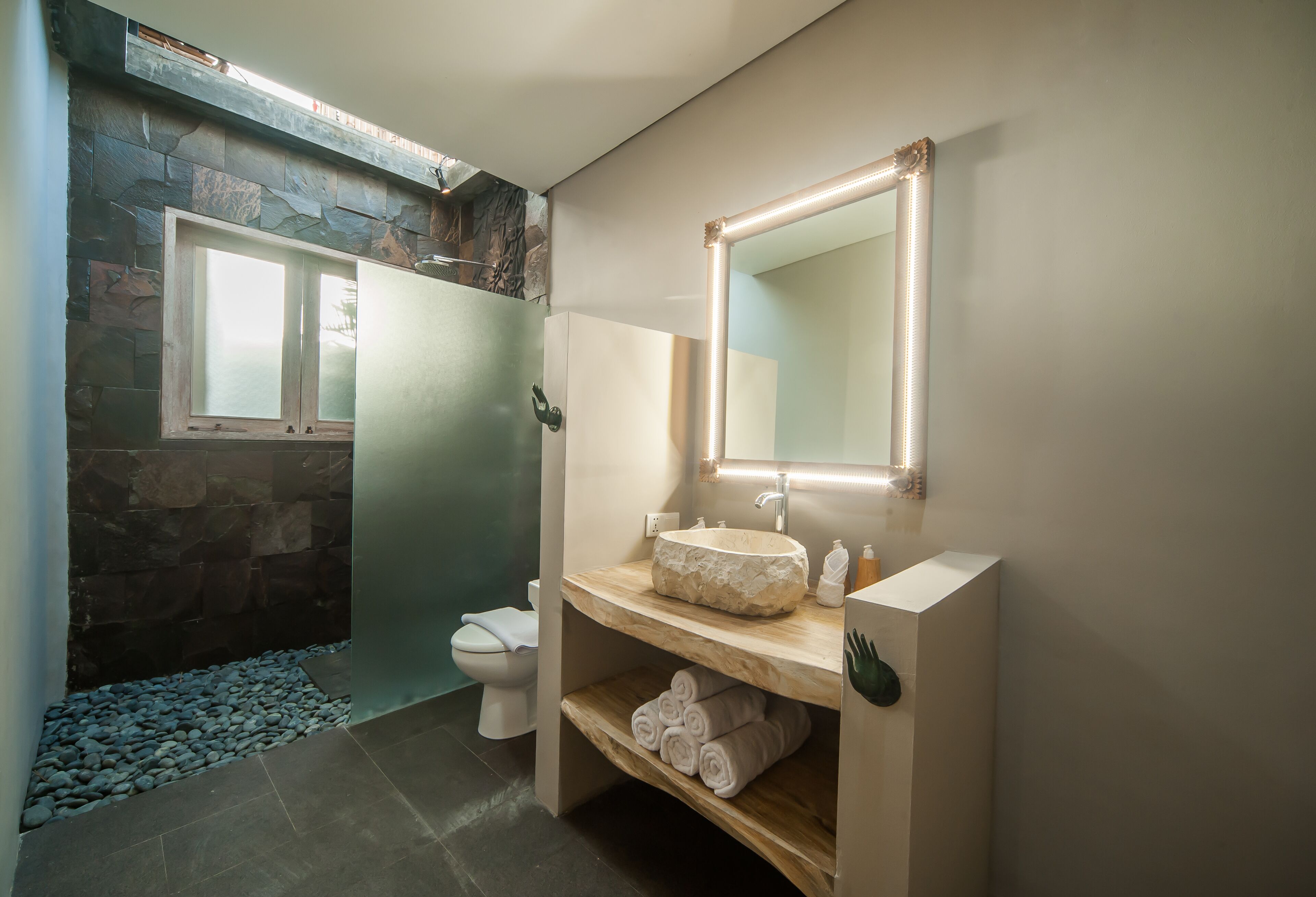 Ein Badezimmer mit Steinwand, Waschtisch aus Holz, Spiegel, Toilette und Ablage mit Handtüchern.